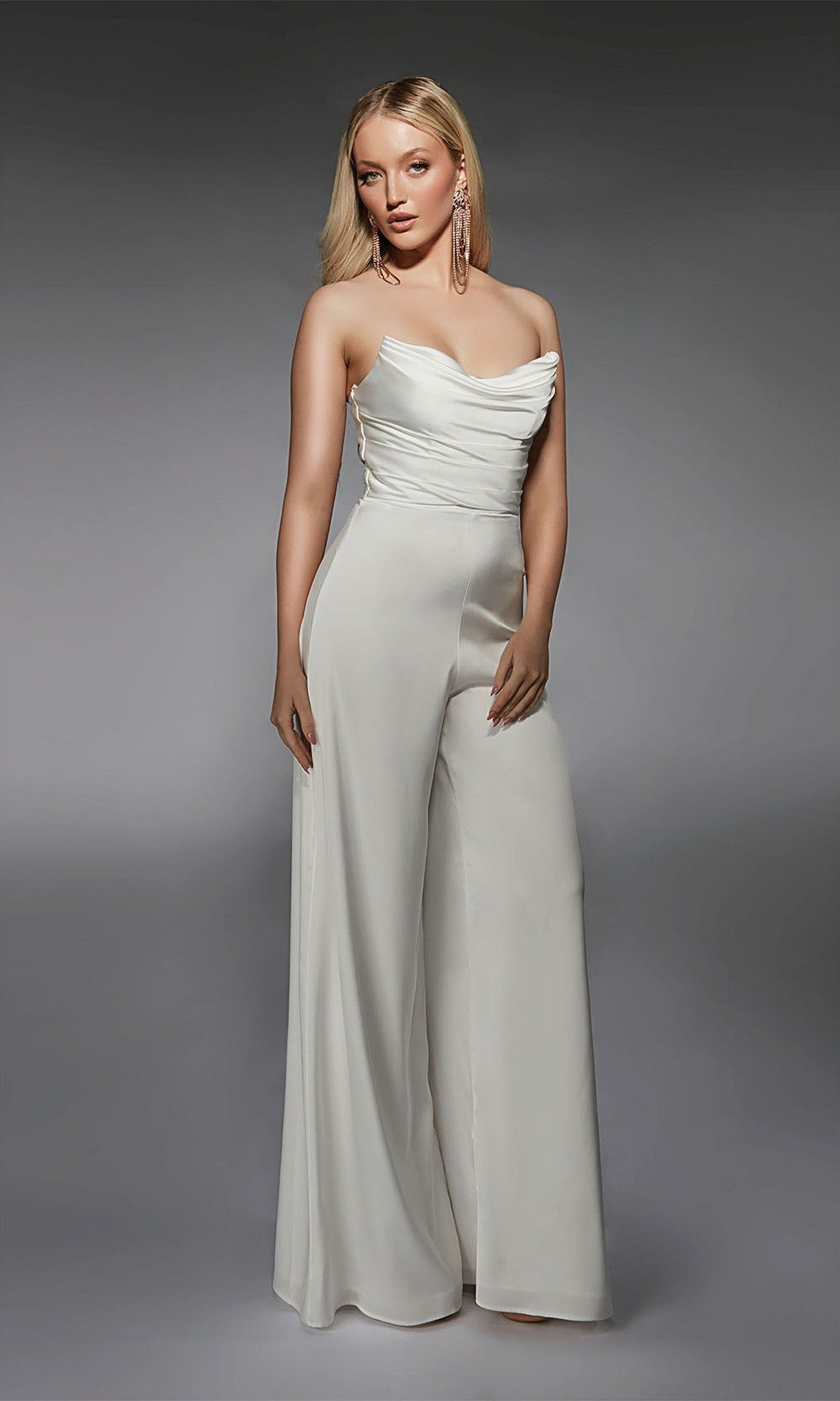 A woman stands against a gray backdrop wearing the Alyce 70071 Long Formal Jumpsuit, a strapless white design with wide-leg pants and statement earrings.